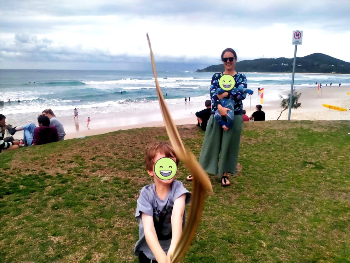 A woman on the Byron Bay foreshore. She has a baby in a carrier on her front, and a young boy waves a long leaf in front of her